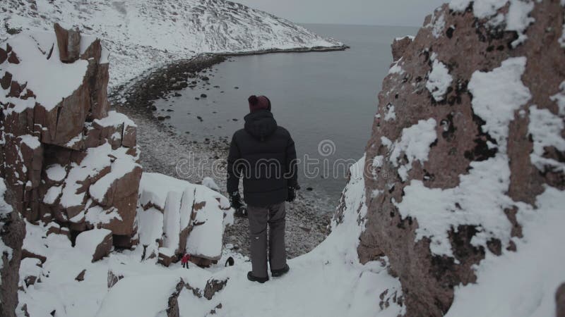 A young man stands in a cliff gorge and admires the view of the dragon eggs beach on the coast of the Barents Sea. Back