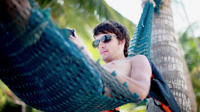 Young man in sunglasses lies on hammock using smartphone on beautiful exotic beach. 1920x1080