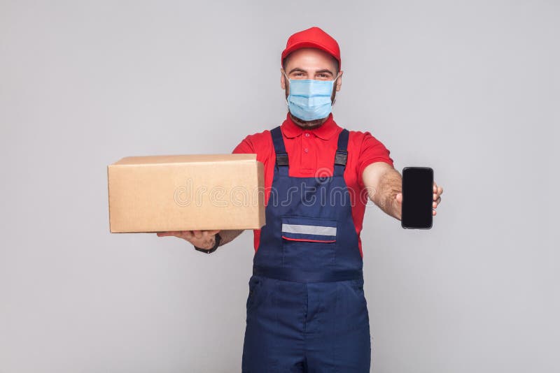 Young man with surgical medical mask in blue uniform and red t-shirt standing, holding cardboard box and showing smart phone