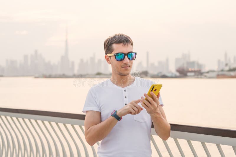Young man is typing on smartphone or using an app on the street against the backdrop of sunset and the Dubai Creek Canal and the royalty free stock images