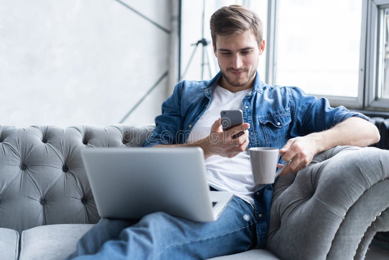 Young man using his smartphone for online banking - sitting on sofa with laptop on leap. stock photography