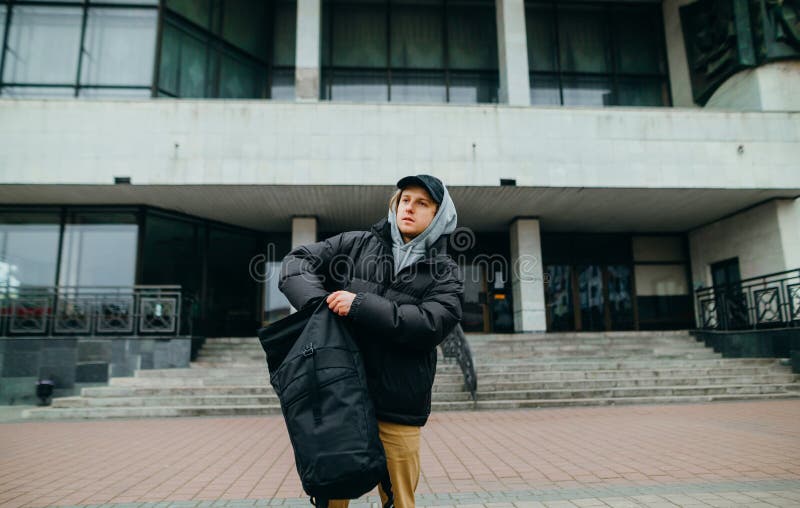 A young man in warm casual clothes stands on the street in the background of the building and pulls things out of his backpack and royalty free stock photo