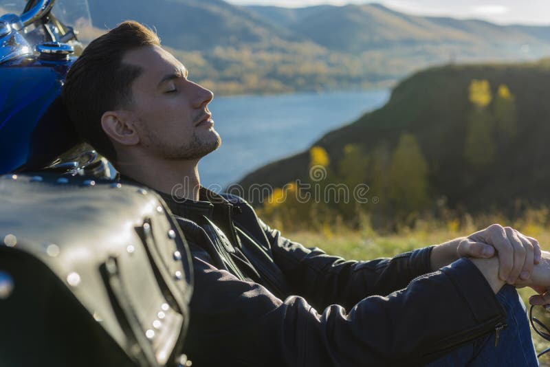 Young man wearing a black leather jacket and jeans sits outdoor stock photo Young man wearing a black leather jacket and jeans sits outdoor stock photo