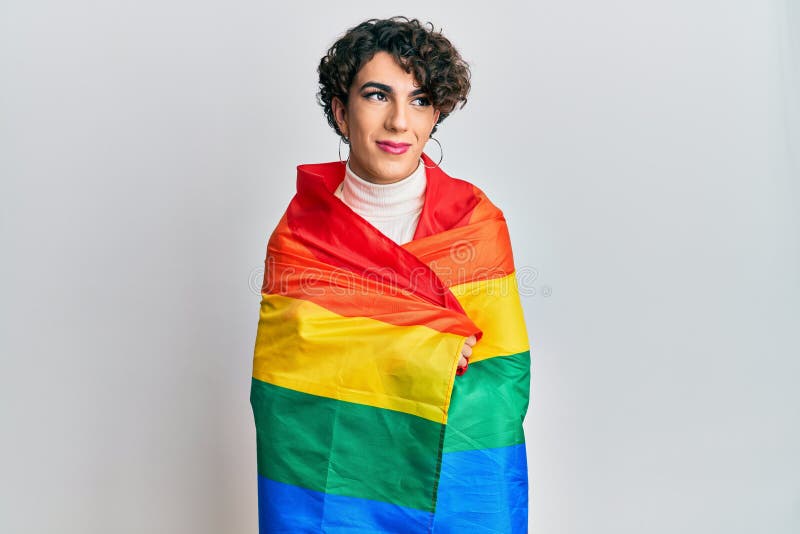 Young man wearing woman make up wrapped in rainbow lgbtq flag smiling looking to the side and staring away thinking