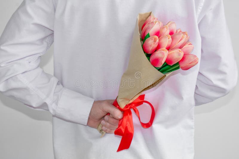 A young man in a white shirt, holding a bouquet of tulips, in his hand behind his back, on a white background. Valentine`s Day