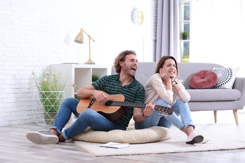 Young man playing acoustic guitar badly for displeased girlfriend in living room.