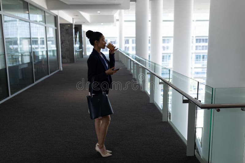 Young mixed-race businesswoman having coffee while using mobile phone in office. Side view of pretty young mixed-race businesswoman having coffee while using stock photo Young mixed-race businesswoman having coffee while using mobile phone in office. Side view of pretty young mixed-race businesswoman having coffee while using stock photo