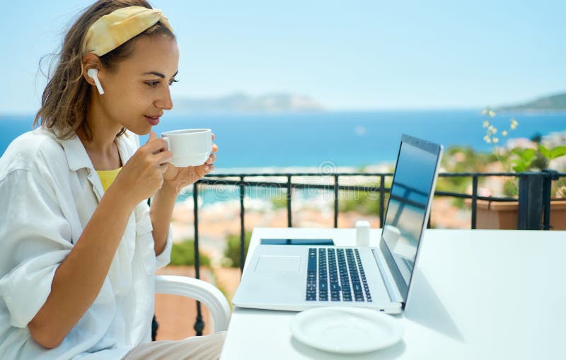 Young mixed race woman in wireless earpods drinking coffee and looking at laptop screen while sitting at balcony resort