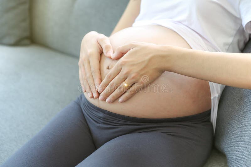 A young mother is making a heart-shaped hand on her stomach. Happy pregnant asian woman with heartshaped by hand in sofa bed stock image A young mother is making a heart-shaped hand on her stomach. Happy pregnant asian woman with heartshaped by hand in sofa bed stock image