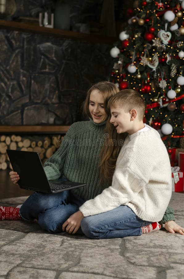 Young mother and teenage son sit near Christmas tree with laptop. Older sister and brother talk to their parents with video call