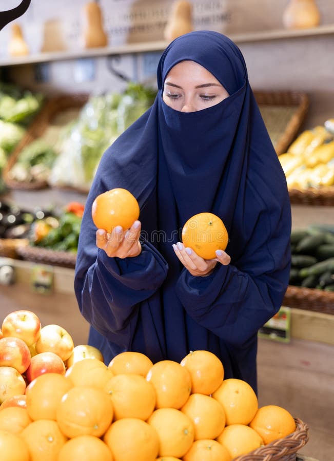 Young Muslim woman in niqab choosing oranges in supermarket. Interested young Muslim woman in traditional islamic dress and niqab making purchases in supermarket