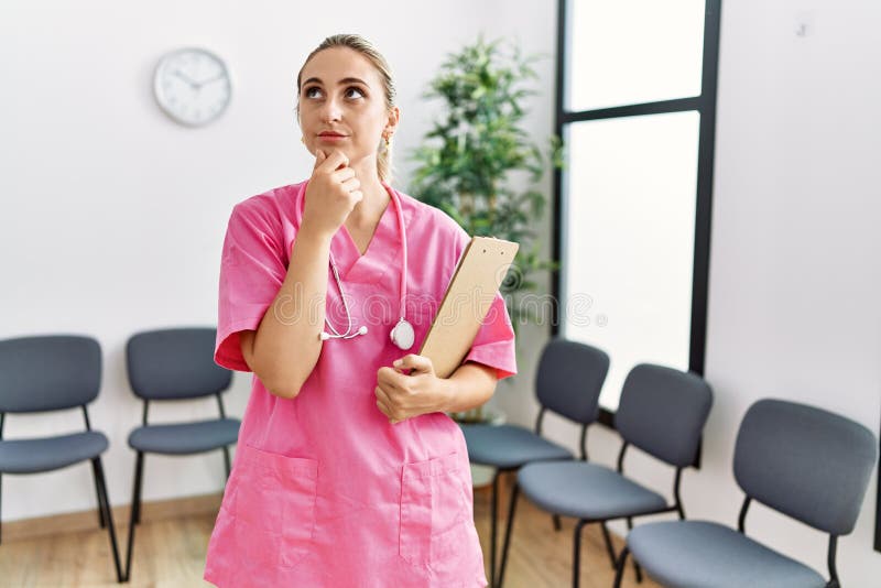 Young nurse woman at medical clinic waiting room serious face thinking about question with hand on chin, thoughtful about