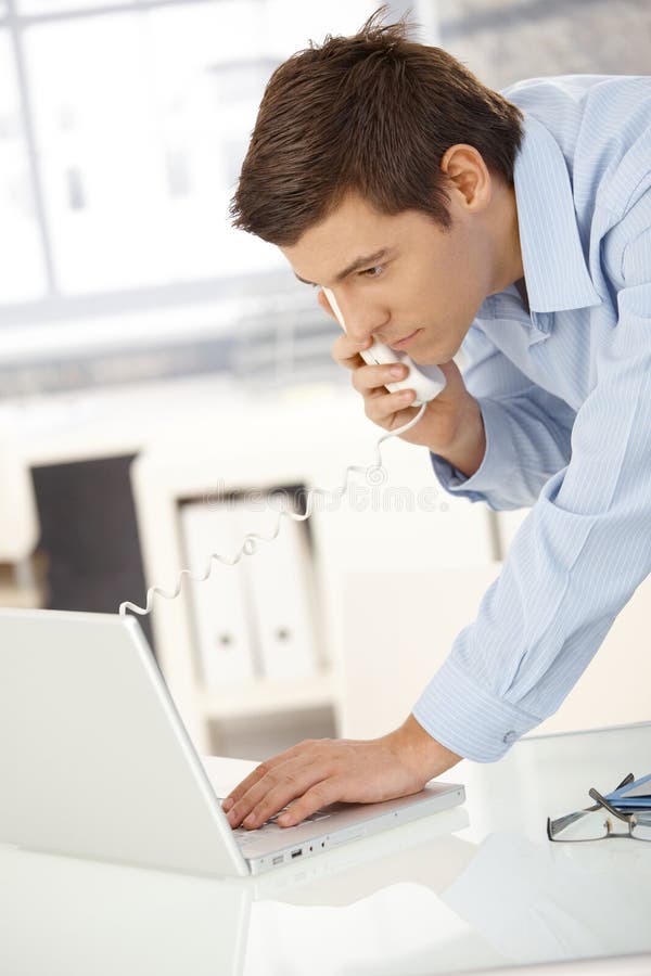 Young office worker man speaking on landline phone using laptop computer, looking at screen. Young office worker man speaking on landline phone using laptop computer, looking at screen.