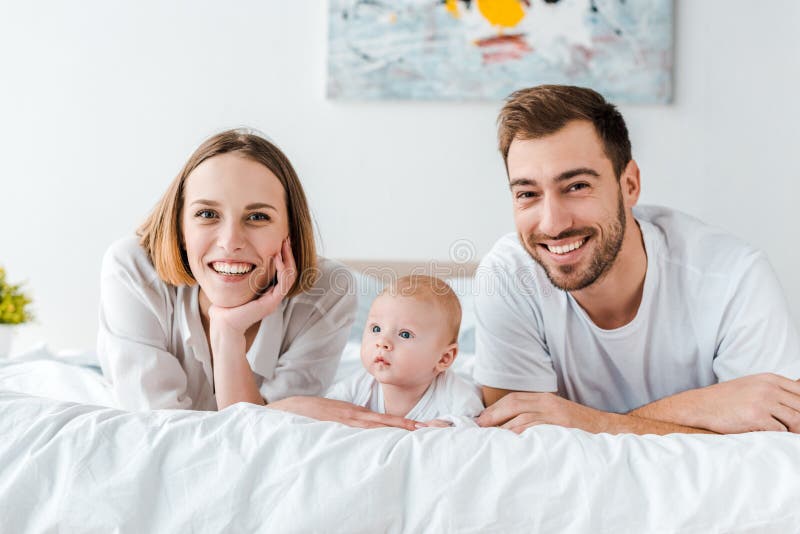 Young Parents with Baby Lying on Bed and Looking at Camera Stock Photo