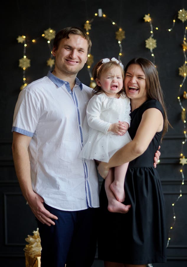 Young parents calm the crying child. The room is festively decorated. Little girl cries on hands at mother. Baby is dressed in a white dress, on the head of her a wreath. Parents look in the camera and smile.