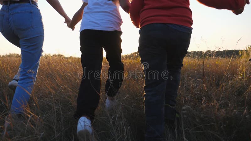 Young parents with kids holding hands of each other and running through grass field at sunset. Mother and father with