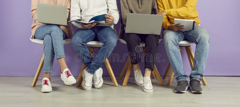 Young people with laptops and notebooks sitting in row on chairs on background of purple wall.