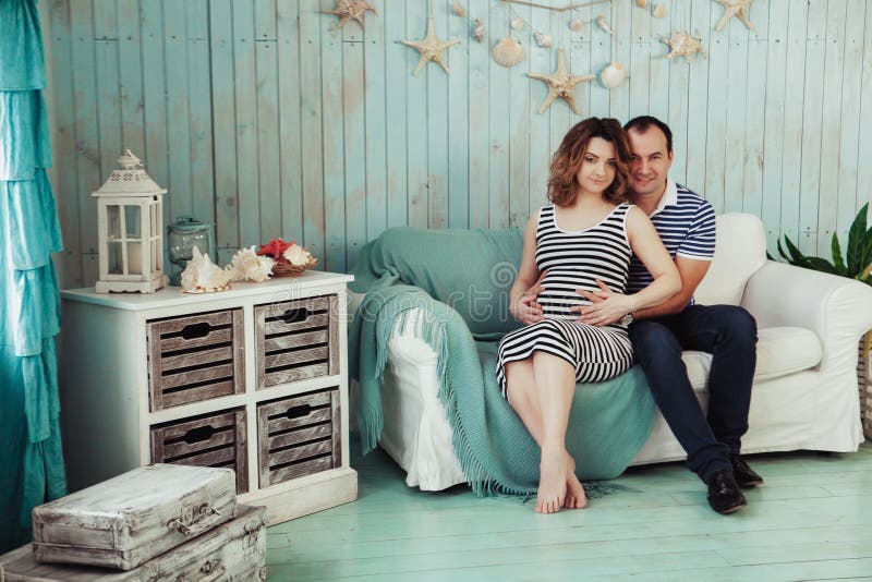 Young pregnant woman with husband on white sofa in blue room. couple dressed in striped clothes. Summer inspiration