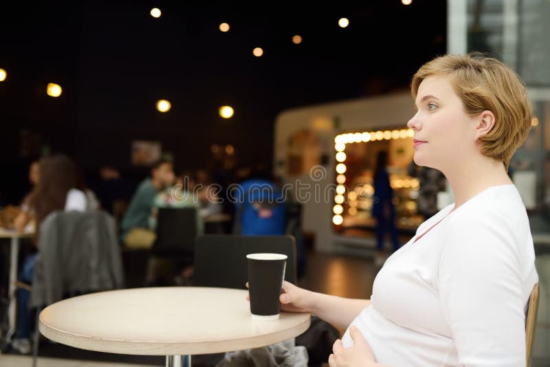 Young pregnant woman resting and drinking coffee at the table of cafe in shopping mall. Nutrition during pregnancy.