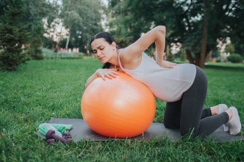 Young pregnant woman suffeer from pain in back. She stand in knees in park. Model lean to orange fitness ball. stock photography Young pregnant woman suffeer from pain in back. She stand in knees in park. Model lean to orange fitness ball. stock photography