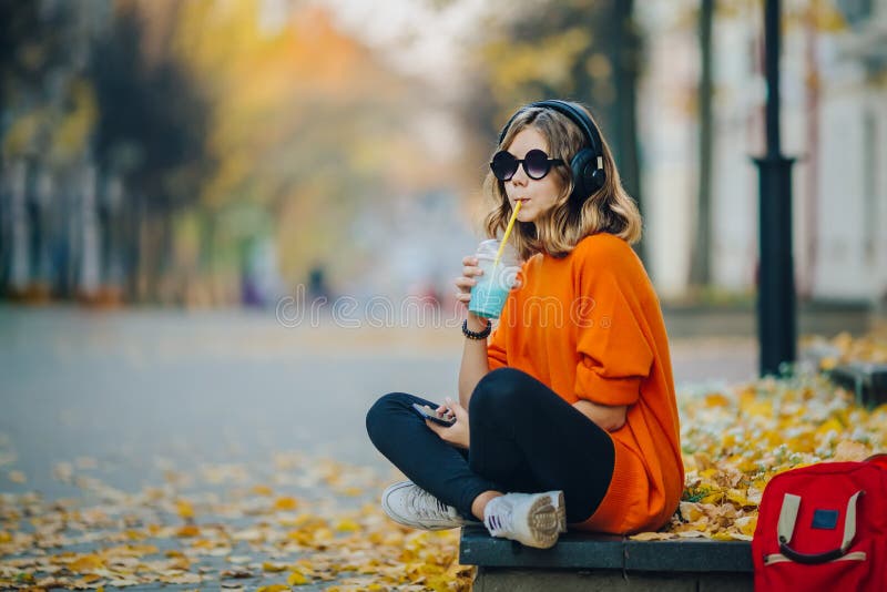 Young pretty hipster teen girl listening music via headphones, sitting on a sidewalk on autumn city street and listening