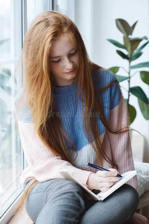 Pretty red haired girl writing notes on notebook with pen, morning light, young woman wearing comfy sweater writing a diary