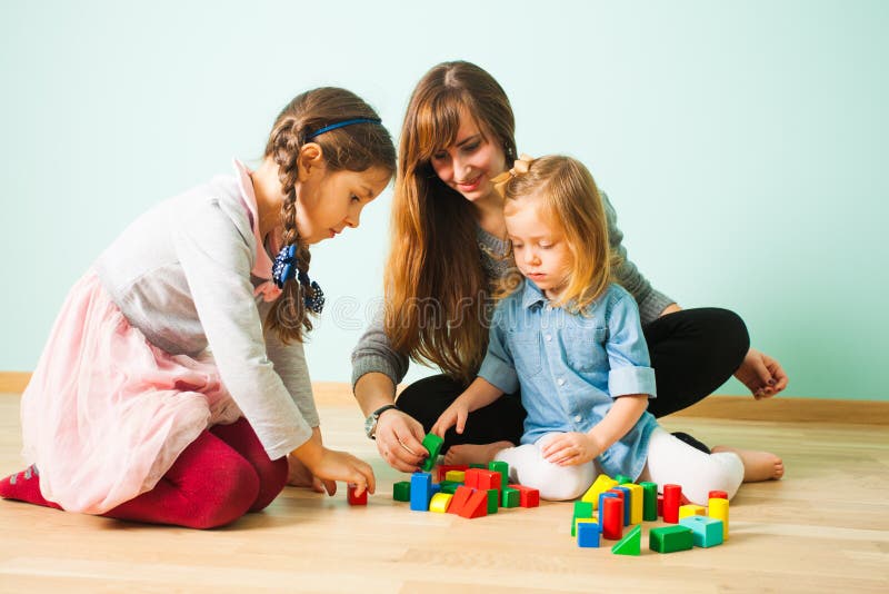 Young pretty smiling nanny playing with two girls sitting on a floor. Woman building towers with two adorable girls while babysitting them. Young pretty smiling nanny playing with two girls sitting on a floor. Woman building towers with two adorable girls while babysitting them.