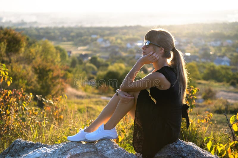 Young pretty woman in black short summer dress sitting on a rock relaxing outdoors at sunset. Fashionable female