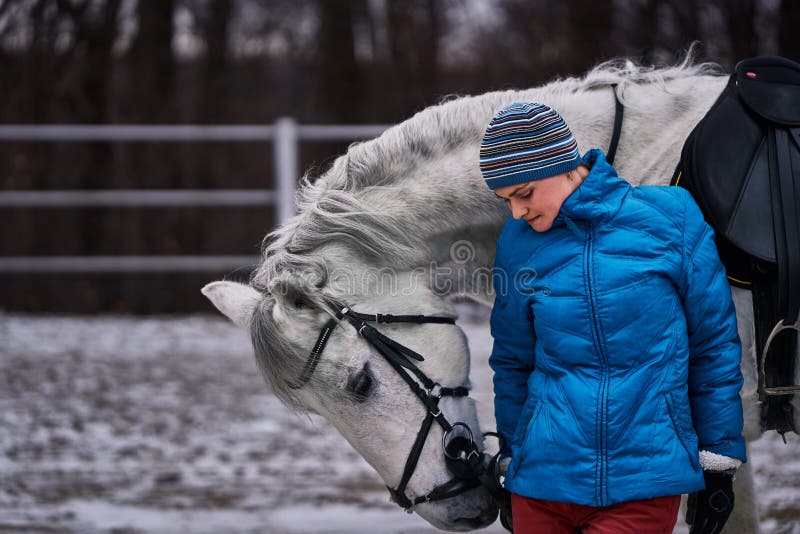 Young pretty woman in blue jacket and sports hat on a walk with a white horse on a winter cloudy day. stock photo Young pretty woman in blue jacket and sports hat on a walk with a white horse on a winter cloudy day. stock photo