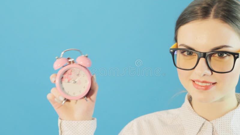 Young Pretty Woman in Glasses and Shirt Holding Pink Clock in Hands on Bright Blue Background in Studio. Time Concept.