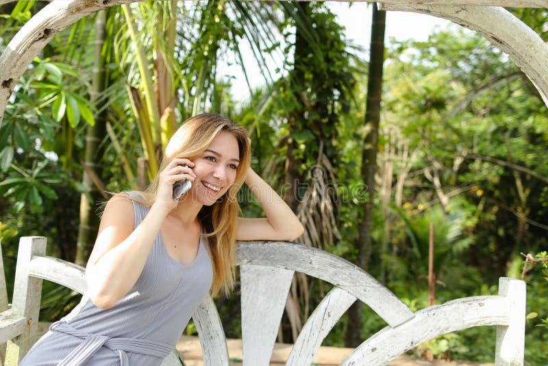 Young pretty woman talking by smartphone with palms in background, sitting on swing. royalty free stock photography Young pretty woman talking by smartphone with palms in background, sitting on swing. royalty free stock photography