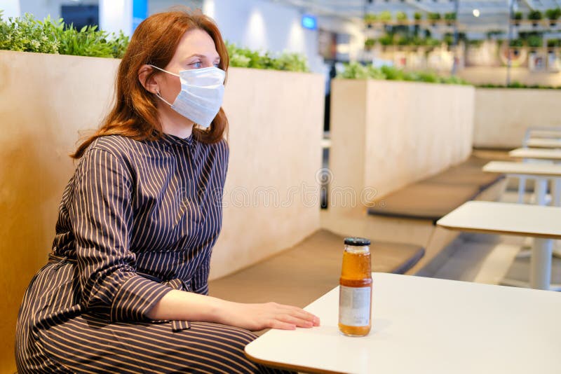 A young red-haired woman in a medical mask sits in an empty cafe during quarantine