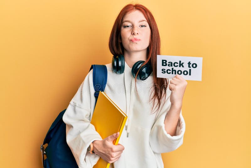 Young red head girl wearing backpack holding back to school banner looking at the camera blowing a kiss being lovely and sexy. love expression