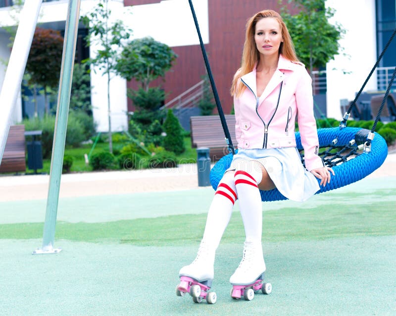 Young redhead woman is swinging on a swing in summer. A fashion trend outfit. Pink jacket and blue summer dress. City park.