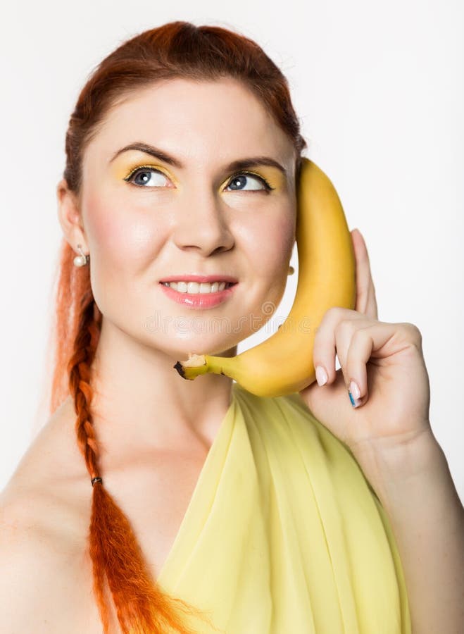 Young redhead woman holding banana near her face like smartphone on a white background.