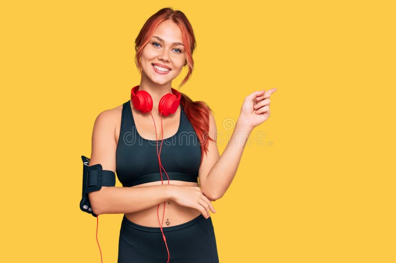 Young redhead woman wearing gym clothes and using headphones smiling happy pointing with hand and finger to the side