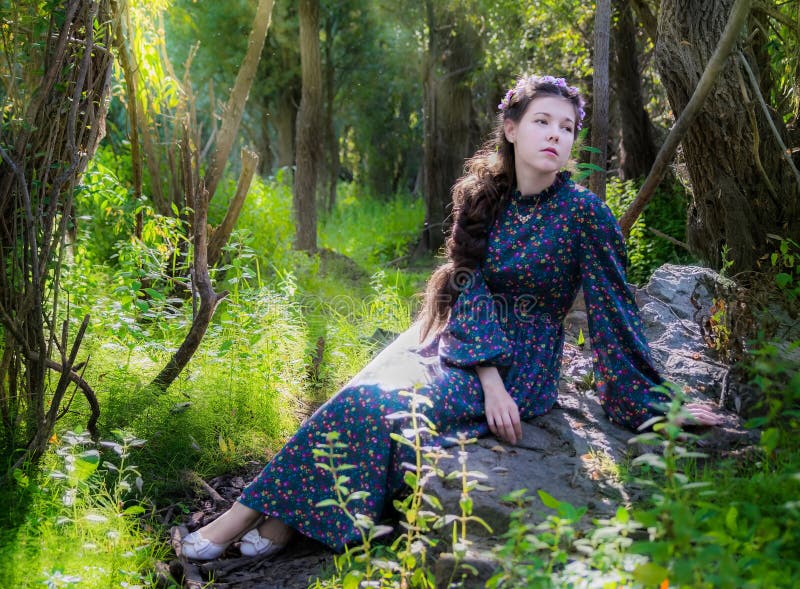 Young sad woman sits on a stone by a tree in a forest