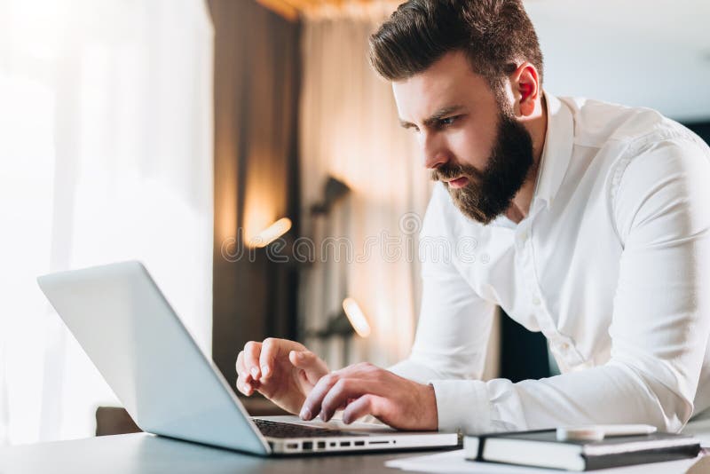 Young man sitting at window and working on laptop. Bearded man sitting at  desktop with smartphone in his hands. Online education. Stock Photo