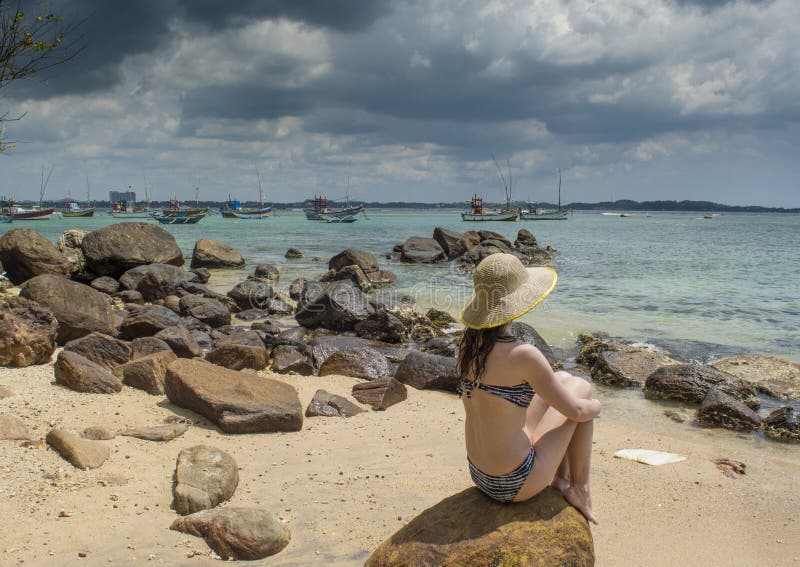 Young sexy beautiful pretty woman in the hat and bikini sitting on huge rock at the beach