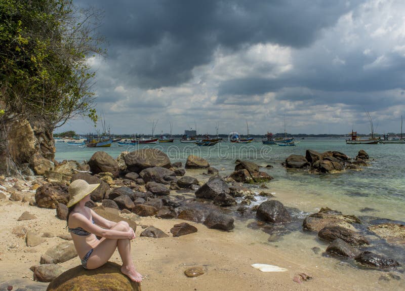 Young sexy beautiful woman in the hat and bikini sitting on huge rock at the beach