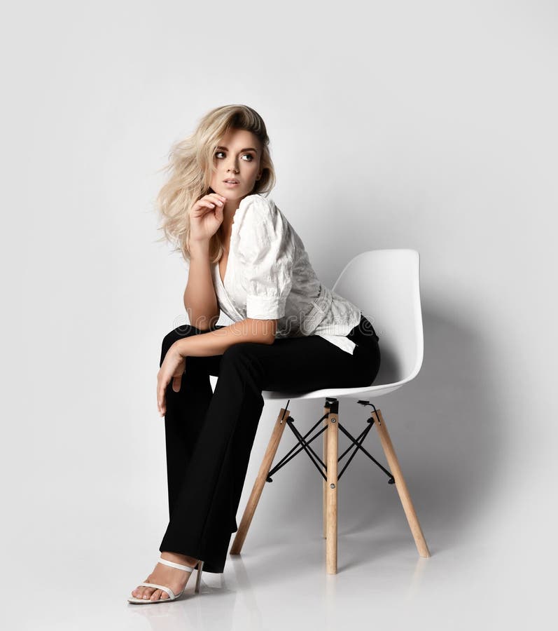 Young sexy blonde slim woman in office pantsuit shirt and pants sits on chair leaning forward and looks aside