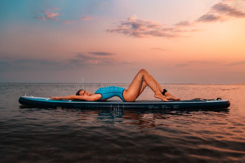 A young sexy caucasian woman in swimsuit lies on a sup board. Sunset in the background. Side view. Copy space. Concept