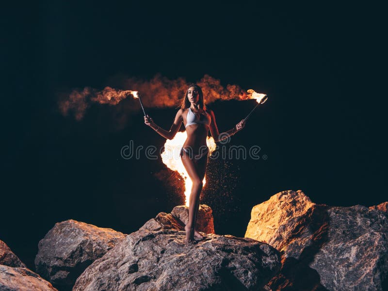 Young sexy woman with a gorgeous slim body in bikini is staying posing at the dark night on the rock with the lit by fire torche