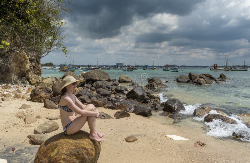 Young sexy woman in the hat and bikini sitting on huge rock at the beach