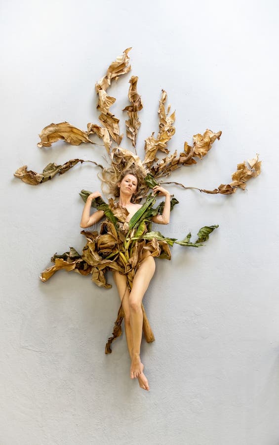 Young sexy woman with long blonde hair artfully covered with dry, withered decorative banana tree leaves lying on the gray studio floor. Copy space