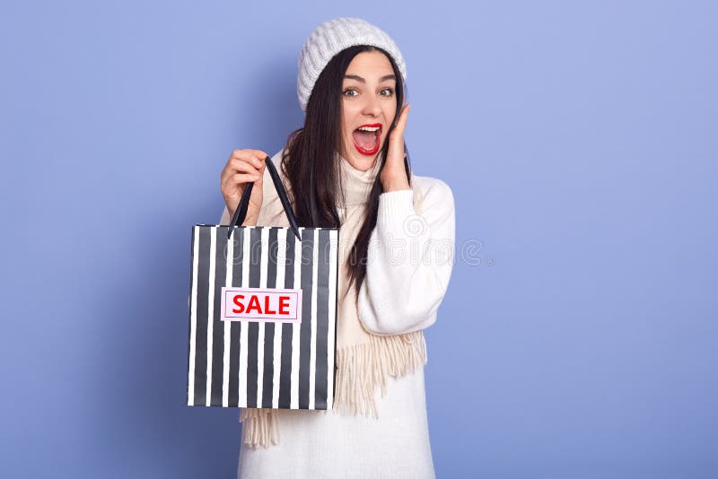 Young shoked lady standing and amazedly looking directly at camera, holding black and white striped shopping bags with inscription