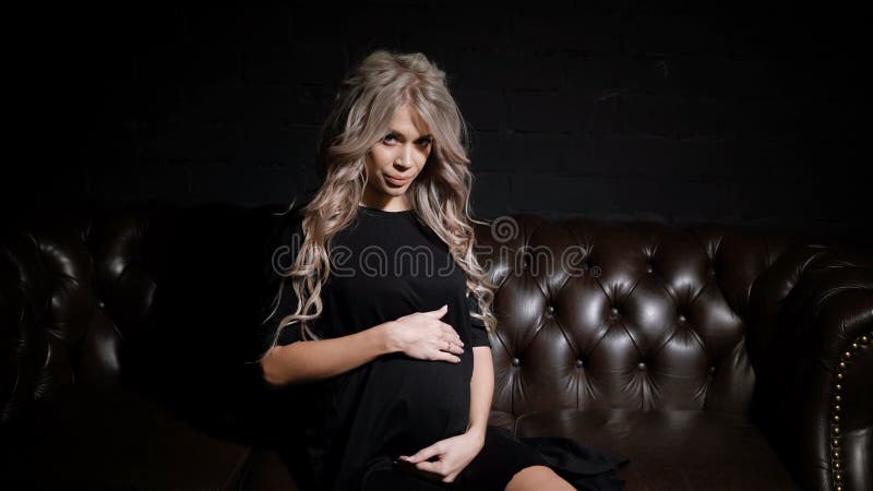 Young single pregnant woman in black dress sitting half-turn in the dark room on the big brown leather sofa looking at