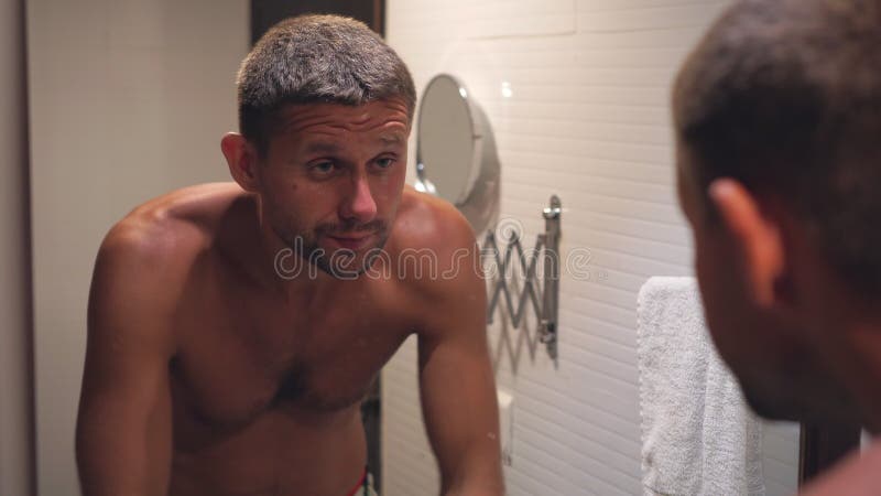 A young sleepy man is washing himself in the bathroom in front of a mirror