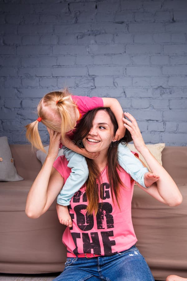 A young slender beautiful mother with her blond daughter on her shoulders play, enjoy and indulge in the teams near the sofa on th royalty free stock photography A young slender beautiful mother with her blond daughter on her shoulders play, enjoy and indulge in the teams near the sofa on th royalty free stock photography