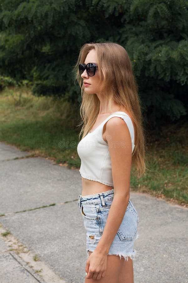 A Young Slender Girl with Long Blond Hair in Short Denim Shorts and a ...
